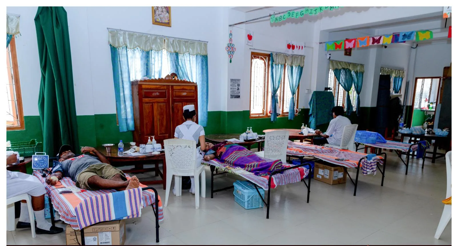 Donors resting on camp beds giving blood, attended by a nurse in a decorated community hall.