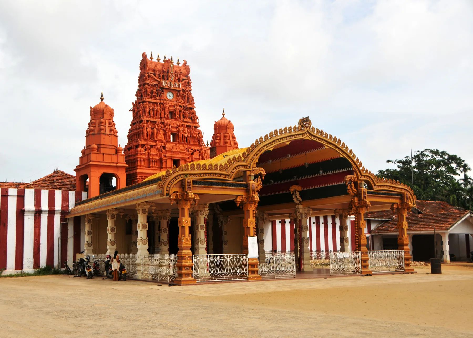 The ornate orange gopuram tower of Nallur Kandaswamy temple against a cloudy sky.
